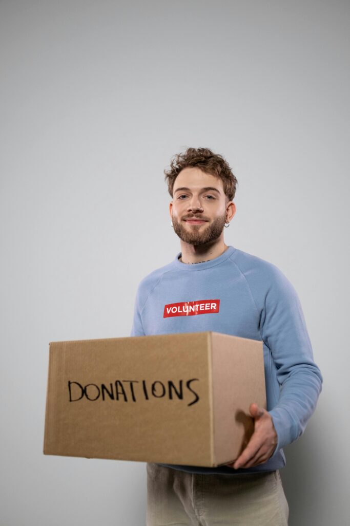 Smiling young man in a volunteer shirt holding a cardboard box labeled donations.